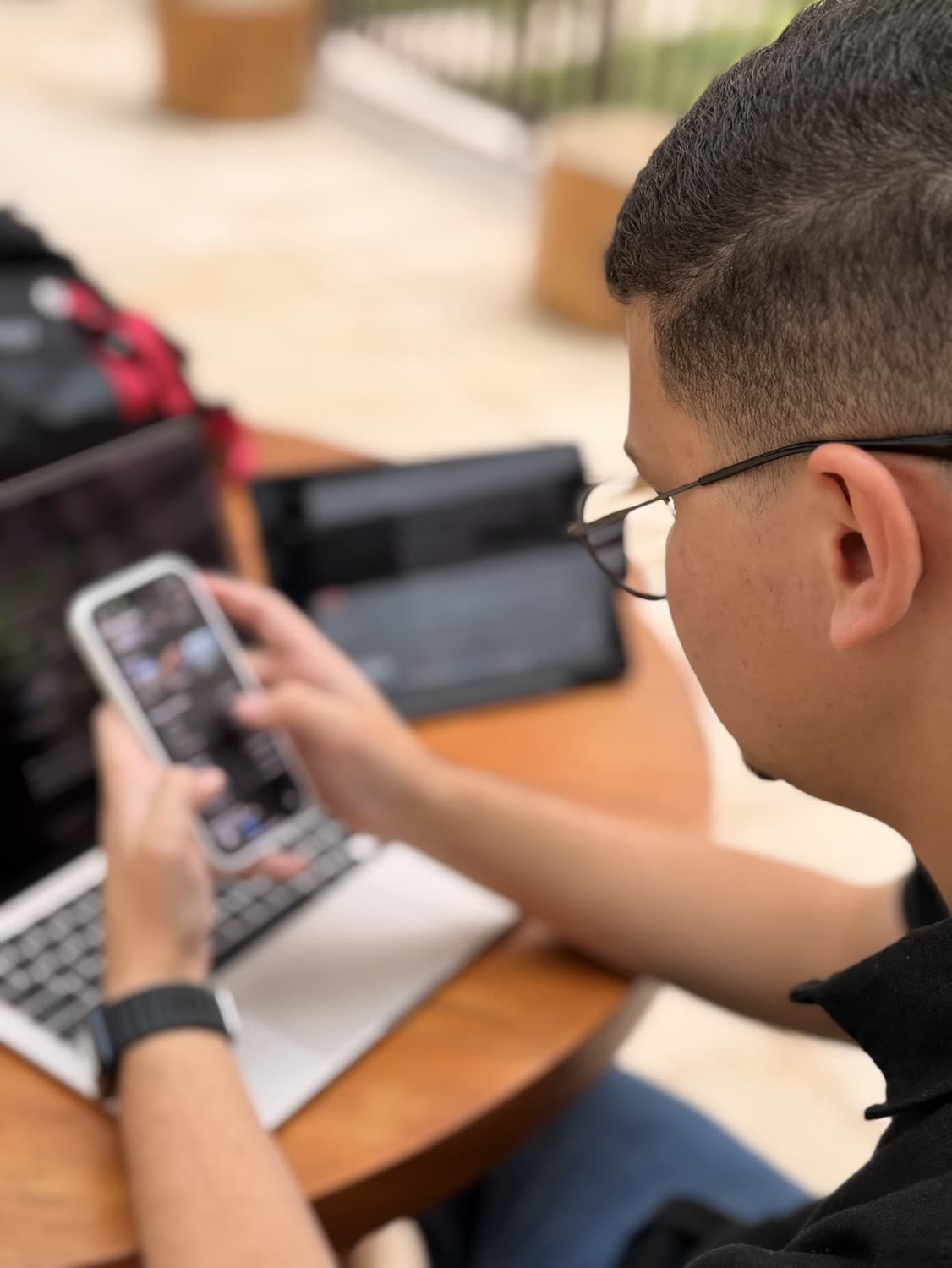 Abdeel visto de lado, de óculos, olhando para o celular, com notebook e tablet sobre a mesa