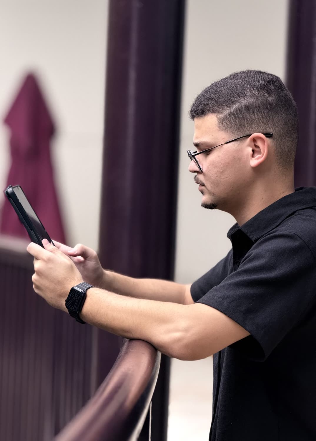 Abdeel de perfil, de óculos e camisa preta, olhando para um celular, apoiado em um corrimão