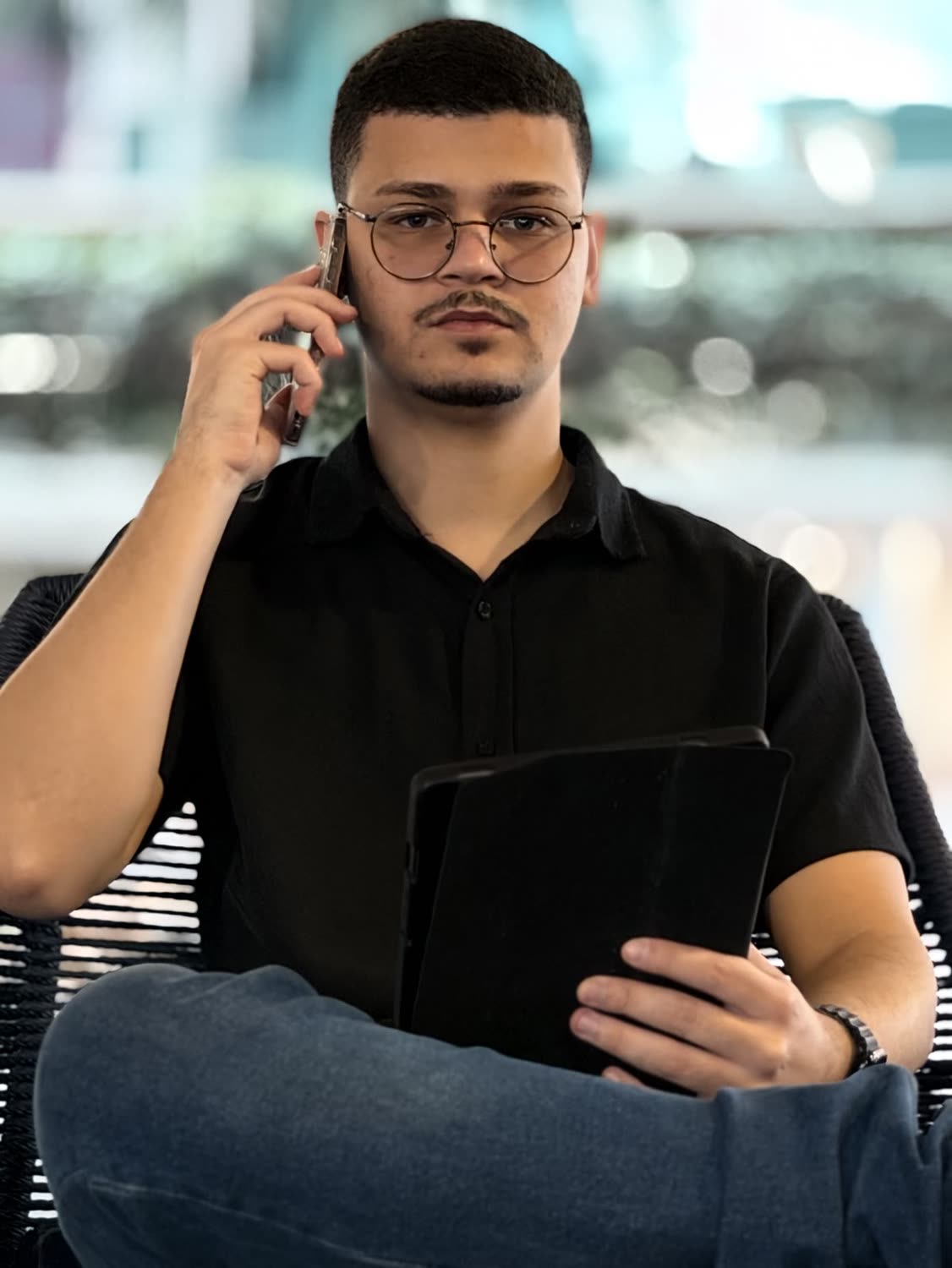 Abdeel sentado, de óculos e camisa preta, ao telefone, com um tablet no colo