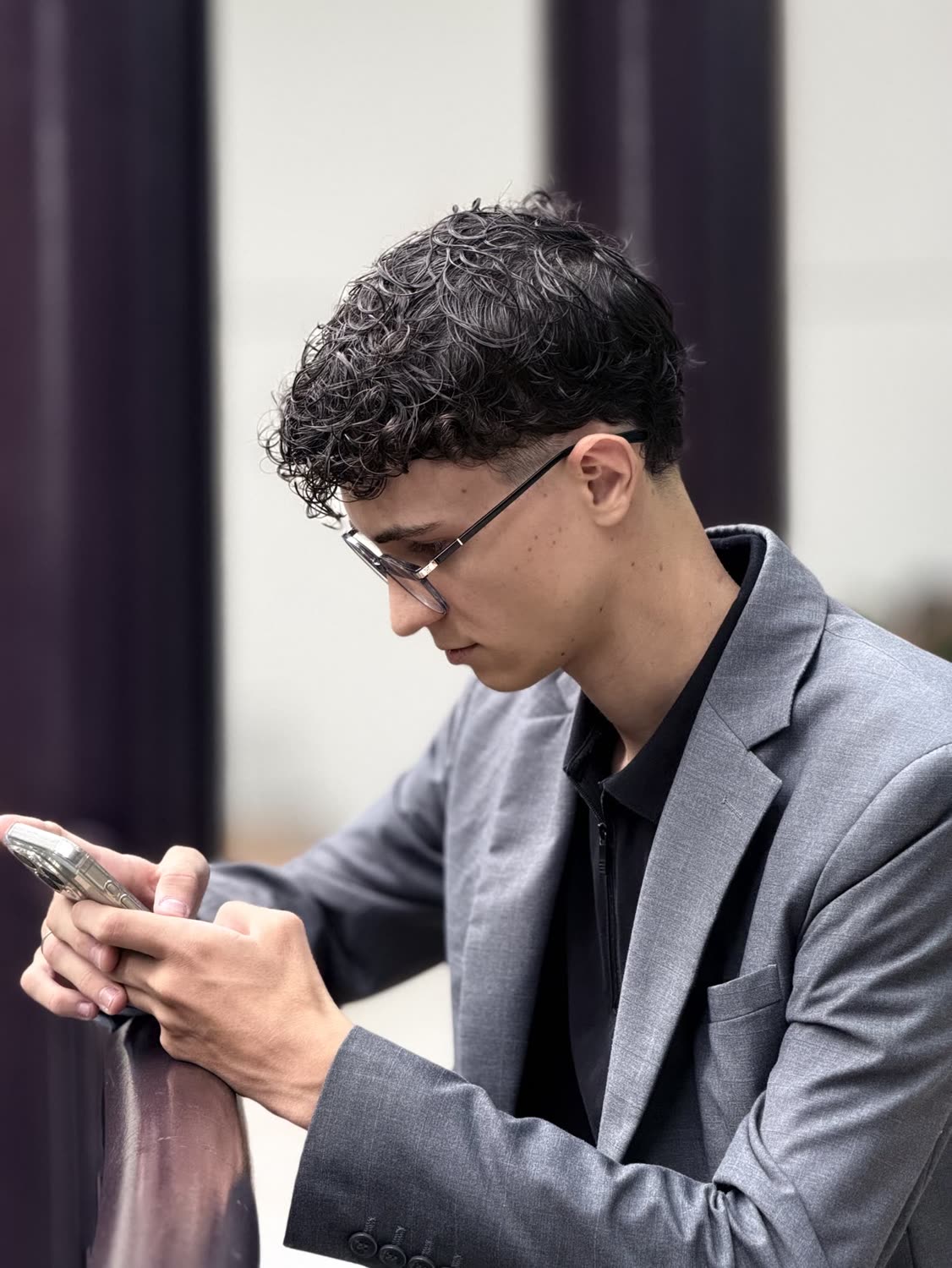 Arthur de perfil, de blazer cinza, digitando no celular, apoiado em um corrimão
