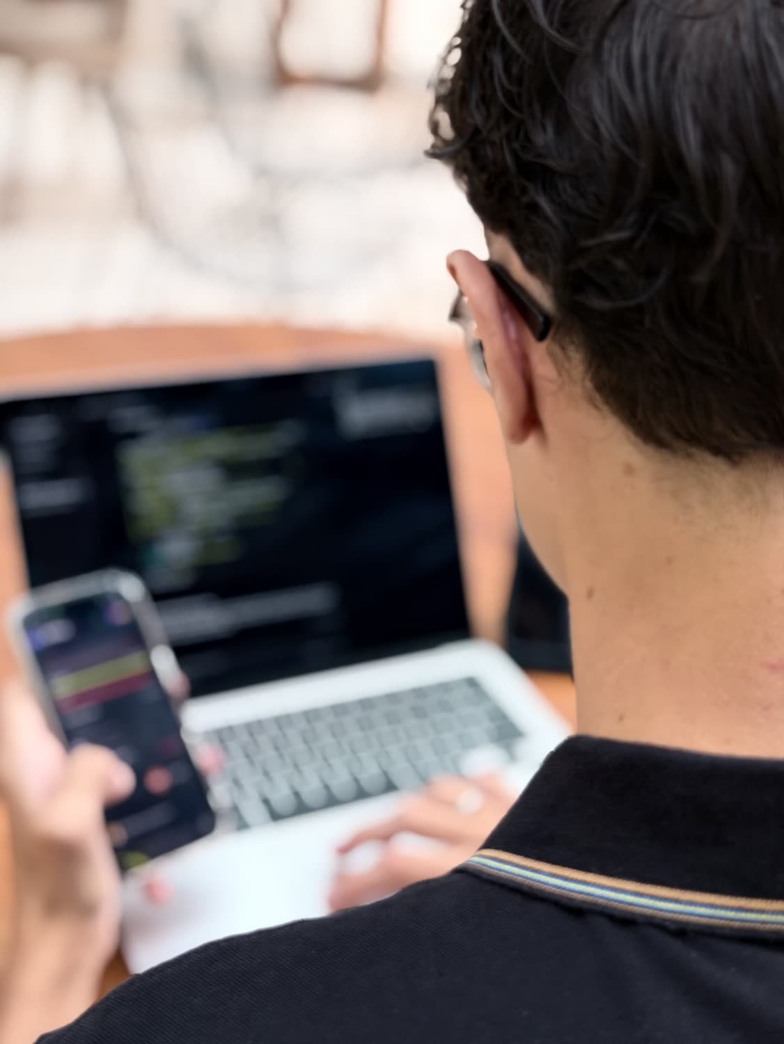 Pessoa de costas diante de um notebook com código na tela, segurando um celular